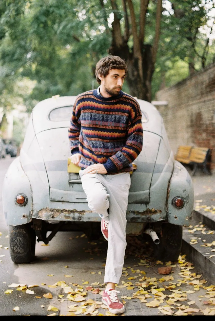 Man in a multicoloured striped knit sweater and light trousers leaning on a vintage car