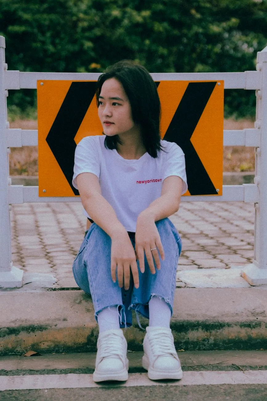 Cô gái mặc áo thun trắng, quần jeans và giày thể thao trắng ngồi bên đường